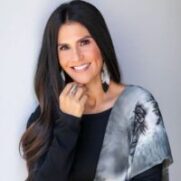 A woman with long dark hair smiles at the camera, wearing a grey blouse with a black pattern and hoop earrings. She poses with her hand on her chin, against a light background.
