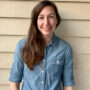 A woman with long brown hair, wearing a denim shirt, smiles warmly while standing against a beige siding background.