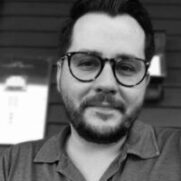 A close-up portrait of a smiling man with a beard and glasses, wearing a gray polo shirt, sitting in front of a blurred background with wooden elements.