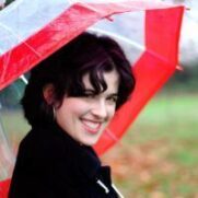 A woman with short dark hair smiling under a red and white transparent umbrella, standing outdoors with greenery in the background.