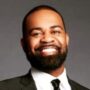 A portrait of a smiling man with a beard, wearing a suit and tie against a gray background.