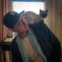 A man wearing a cap and glasses looks thoughtfully to the side with a Siamese cat perched on his shoulder. They are indoors, possibly in a living room.