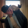 A man wearing a cap and glasses looks thoughtfully to the side with a Siamese cat perched on his shoulder. They are indoors, possibly in a living room.