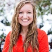 A smiling young woman with wavy blonde hair wearing a bright red shirt, standing in front of a snowy background.