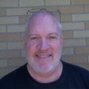 A smiling middle-aged man with glasses resting on his head, wearing a black T-shirt, against a brick wall background.