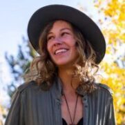A young woman with curly hair wearing a black hat and green shirt, smiling and looking away, surrounded by autumn leaves.