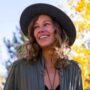 A young woman with curly hair wearing a black hat and green shirt, smiling and looking away, surrounded by autumn leaves.