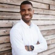 A smiling man with crossed arms wearing a white shirt stands in front of a wooden slat background. He appears confident and approachable.