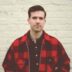 A man in a red and black plaid shirt standing in front of a beige wall, looking directly at the camera with a neutral expression.