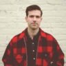 A man in a red and black plaid shirt standing in front of a beige wall, looking directly at the camera with a neutral expression.