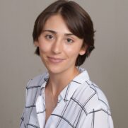 A professional headshot of a smiling woman with short brown hair, wearing a white and black checkered shirt, against a plain light gray background.