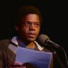 A man with glasses and a striped sweater reads from a paper at a microphone on a dimly lit stage.