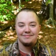 A smiling person with short hair wearing a camouflage jacket, standing in a forest with greenery in the background.