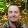 A smiling person with short hair wearing a camouflage jacket, standing in a forest with greenery in the background.