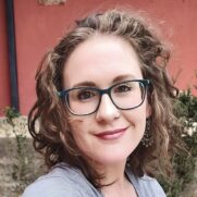 A woman with curly hair and glasses smiling at the camera, wearing a grey top and earrings, with a red door and greenery in the background.