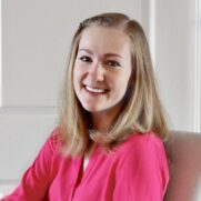 A smiling young woman with blonde hair wearing a bright pink blouse, seated indoors with a soft-focus, neutral background.