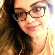 A woman with glasses and dark wavy hair, wearing a yellow blouse, looks directly at the camera with a gentle smile.