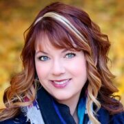 A woman with blue eyes and curly brown hair with blonde highlights smiles warmly. She wears a scarf and stands against a blurred background of autumn leaves.