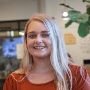 A smiling young woman with long blonde hair, wearing a burnt orange top, standing in an office environment with a blurred background featuring a sign and a plant.