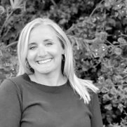 Black and white portrait of a smiling woman with blond hair, wearing earrings and a dark sweater, standing in front of leafy plants.