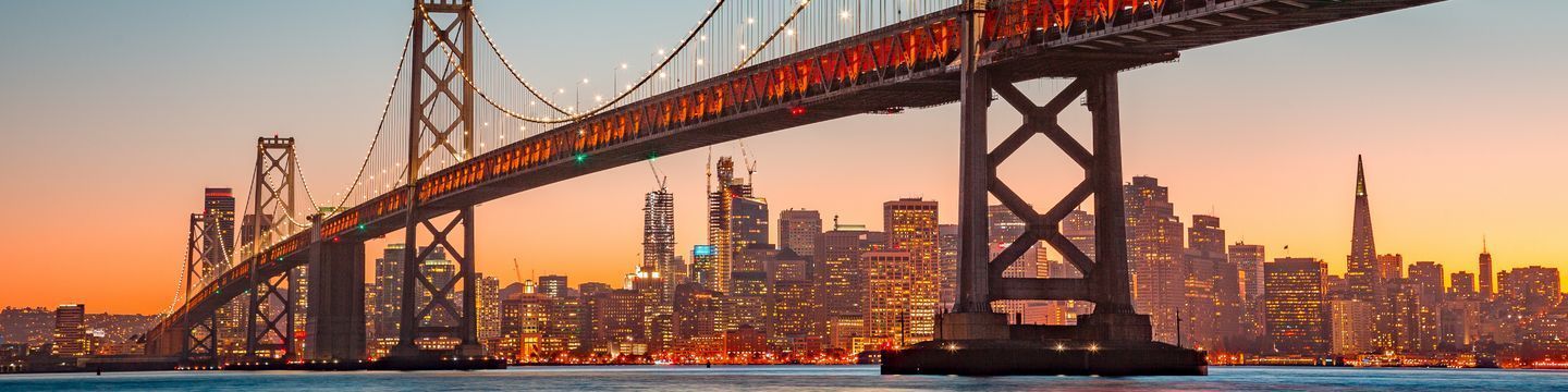 Panoramic view of a lit-up suspension bridge at twilight with a city skyline in the background under an orange sky, embodying the essence of California destinations.