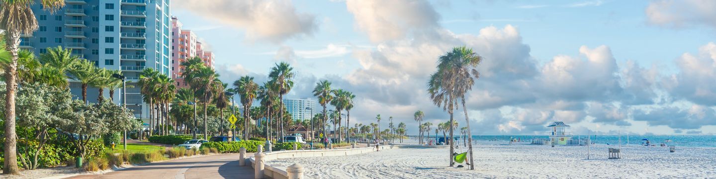 A panoramic view of a beachside promenade lined with palm trees, adjacent to a wide sandy beach typical of Florida beaches, with a clear blue sky, colorful beachfront buildings, and a lifeguard