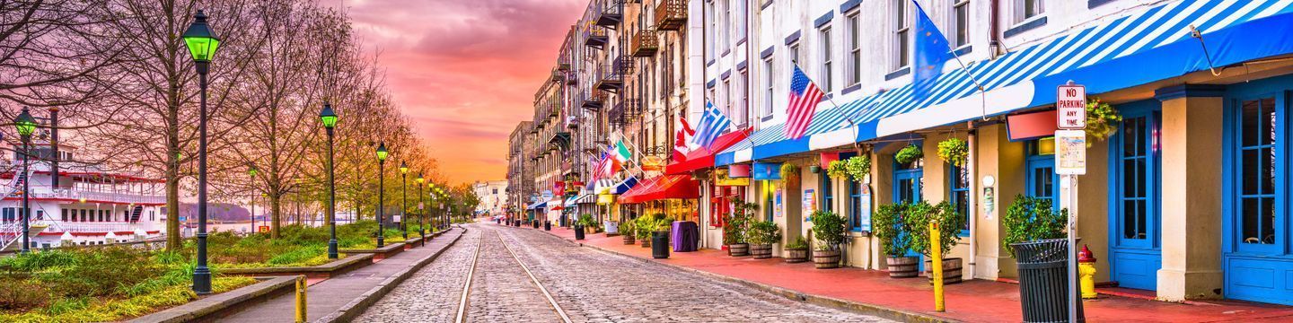 A vibrant panoramic view of a city street in Georgia featuring colorful buildings and a cobblestone pavement, leading towards a river with a moored boat, under a colorful sky.