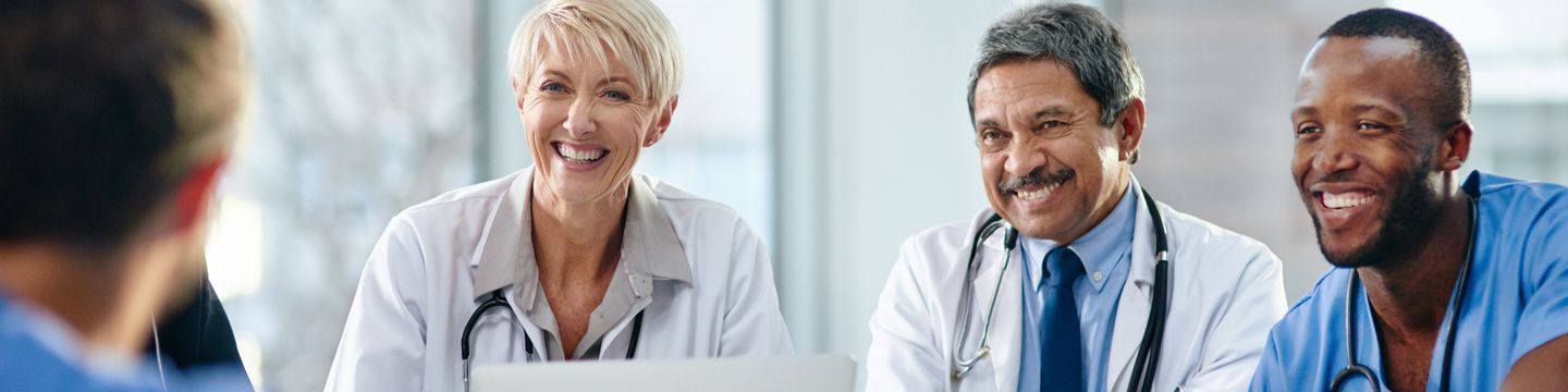 Three healthcare professionals—a white woman, a middle eastern man, and a black man with accreditation—smiling and engaging in a discussion with a fourth person, possibly a patient, not visible in the image