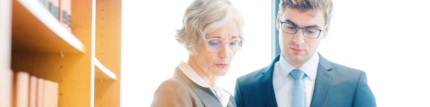 A panoramic image featuring two professionals in a well-lit office: an elderly woman with glasses reading a document related to criminal justice and a young man in a suit looking at the same document.