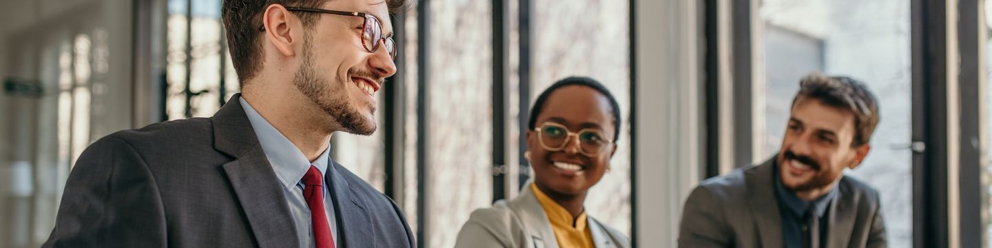 Three professionals in a sunlit office, smiling and engaging in a lively discussion. One woman and two men, dressed in business attire, are celebrating one year of collaboration.