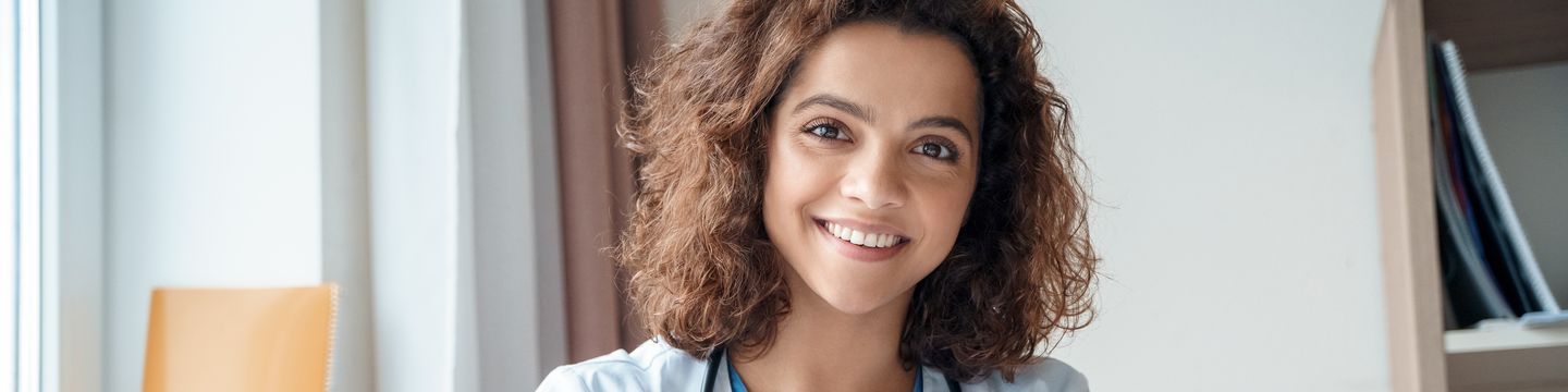 A cheerful female doctor with curly hair wearing a lab coat, sitting in a bright office, smiles confidently at the camera.