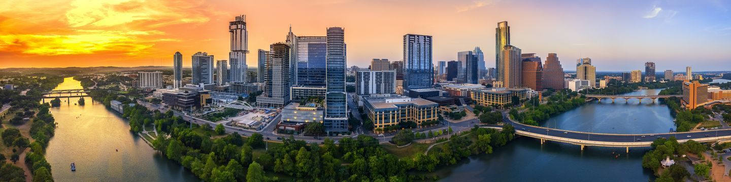 Panoramic aerial view of a vibrant Texas city skyline at sunset, featuring modern skyscrapers, a river flowing through the center, and bridges connecting both sides.