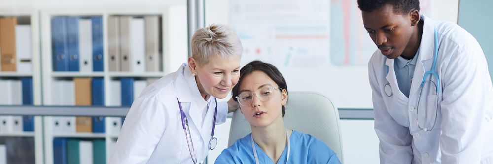 Two doctors, one male and one female, assisting a female patient seated in a chair, in a clinical setting with medical charts in the background.