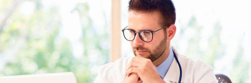 A focused male doctor with glasses and a stethoscope around his neck, sitting at a desk looking at a laptop in a bright office.