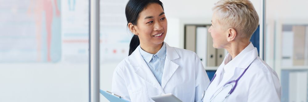 Two female healthcare professionals in white lab coats are conversing; one holds a clipboard while the other listens attentively in a clinical setting.