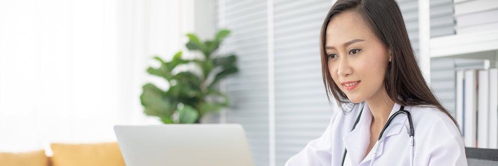 A female doctor in a white lab coat with a stethoscope around her neck, working intently on a laptop in a bright office setting.