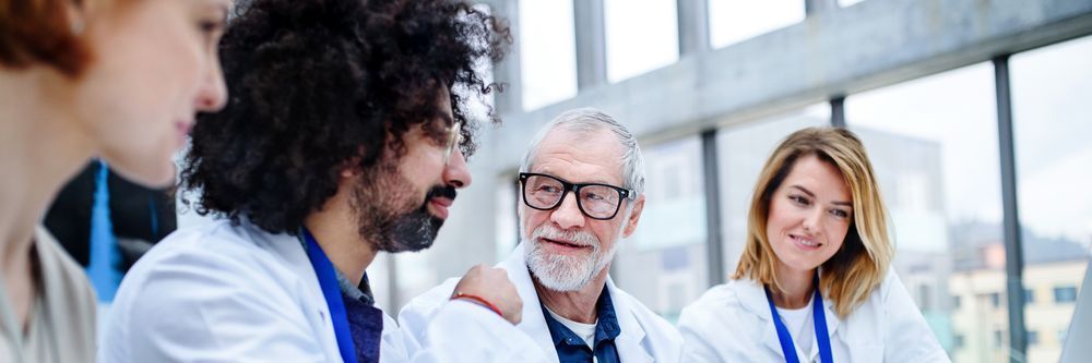 A group of four medical professionals, wearing white coats and ID badges, engaging in a discussion in a bright, modern office setting.