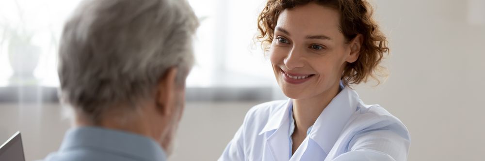 A female Doctor of Social Work with curly hair and a lab coat smiling at an elderly male patient during a consultation in a bright, modern office.