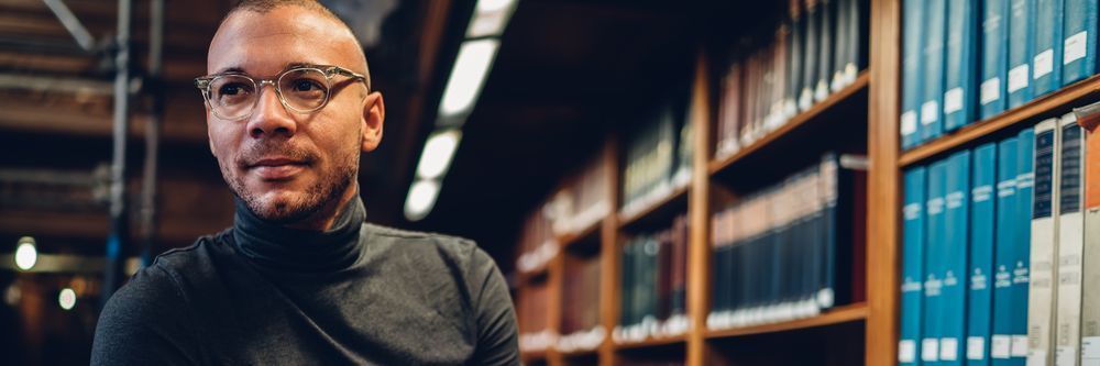 A confident man wearing glasses and a black turtleneck smiles subtly while standing in a library with rows of books behind him.