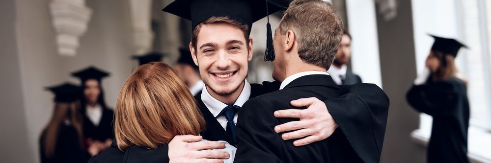 A young graduate in cap and gown hugs two adults, smiling joyfully, during a graduation ceremony.