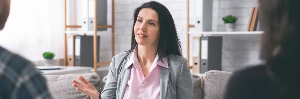 A professional woman in a pink shirt and blazer gestures while conversing with two unseen individuals in a modern living room setting.