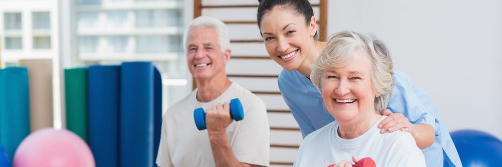 Two elderly individuals exercising with dumbbells in a gym with the assistance of a smiling female instructor.