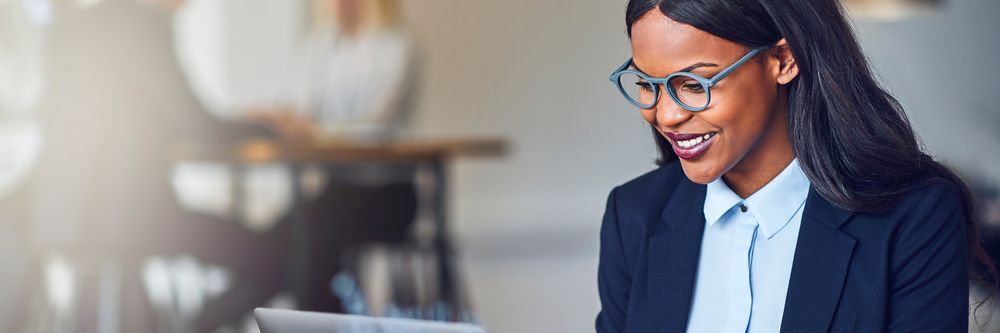 A professional Black woman with glasses smiles while working on a laptop in a modern office environment, with colleagues in the background.