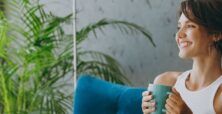 A woman smiles contentedly while holding a mug, sitting on a couch with blue pillows, next to a window with lush green plants.