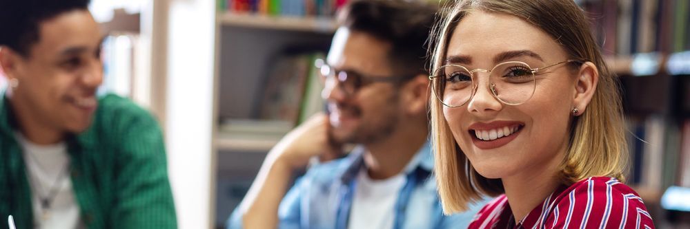 A young woman with glasses, smiling broadly, foregrounded in a library with two male colleagues conversing softly in the background.