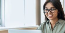 A smiling woman with glasses using a laptop at a desk by a window with a potted plant to the side.