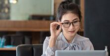 A young Asian woman in business attire adjusts her glasses while looking at a laptop in a modern office setting.