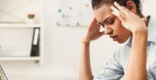 A distressed woman sits at a desk, touching her temples and looking at a laptop screen in a home office environment.