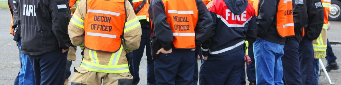 Group of emergency responders, including fire and medical officers, standing in a circle. They are wearing high-visibility vests and jackets labelled "FIRE," "DECON OFFICER," and "MEDICAL OFFICER," among others. They appear to be in discussion or briefing.
