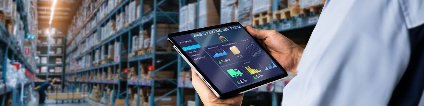 A person holding a tablet in a warehouse with shelves of boxes. The tablet screen displays various charts and data for warehouse inventory management, including graphs and percentage figures. The background shows a well-lit, organized storage area.